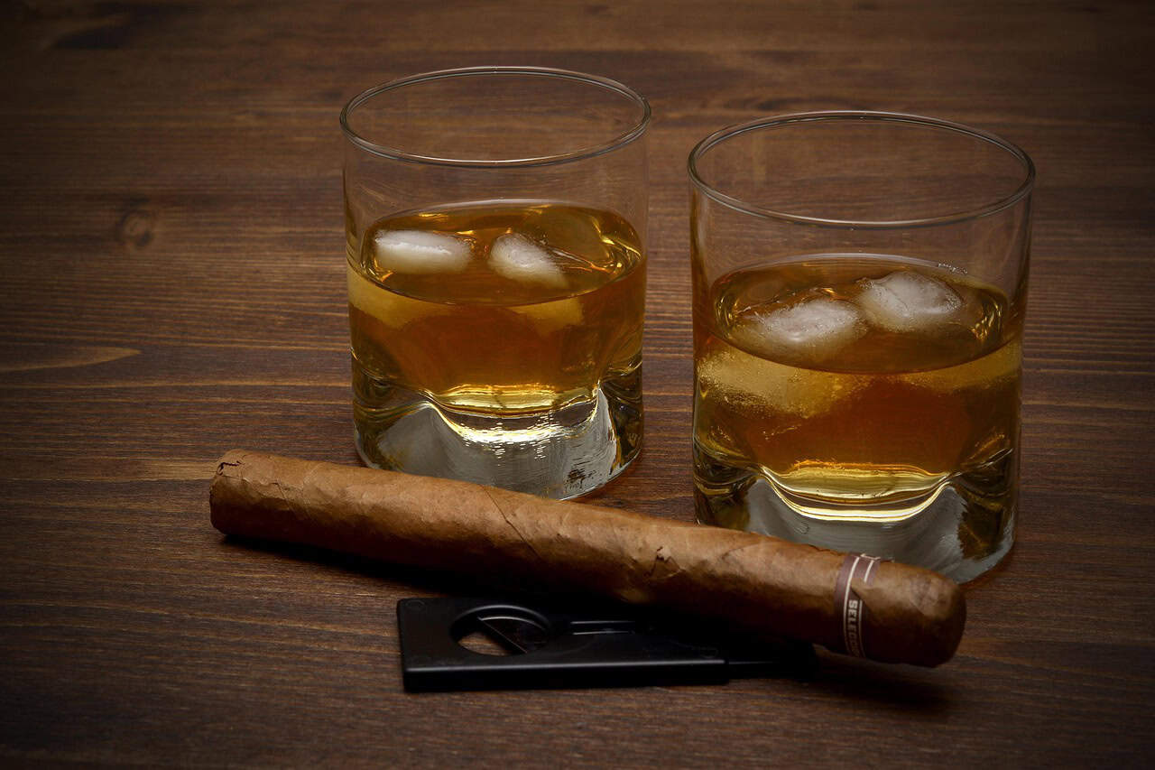 Two glasses of non alcoholic whiskey with ice cubes sit on a wooden surface next to a cigar and a cigar cutter.