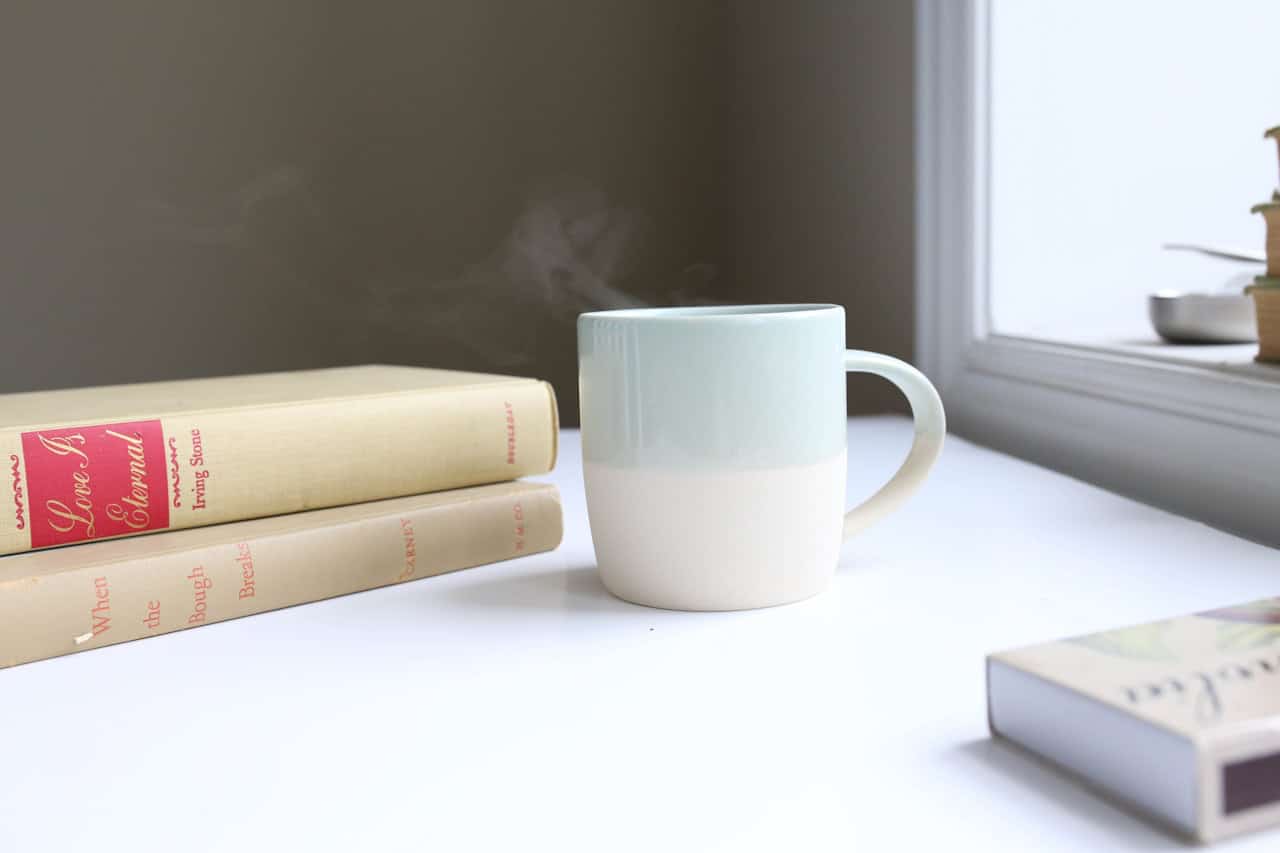 A steaming white and light blue mug sits on a white table beside two stacked hardcover books and a window, inspiring cozy book club food ideas for your next gathering.