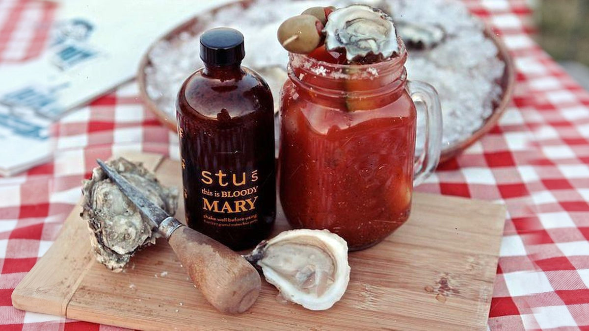 Fresh shucked oysters with an oyster knife, a Bloody Mary with an oyster garnish, and a bottle of Stu's Bloody Mary seasoning on a checkered tablecloth