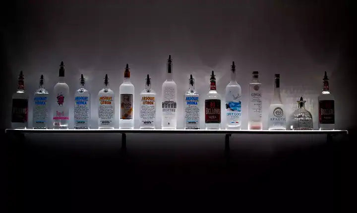 A row of various vodka bottles, including brands like Absolut, Belvedere, Grey Goose, and Ciroc—perfect vodkas for Bloody Mary—displayed on a backlit shelf against a plain wall.