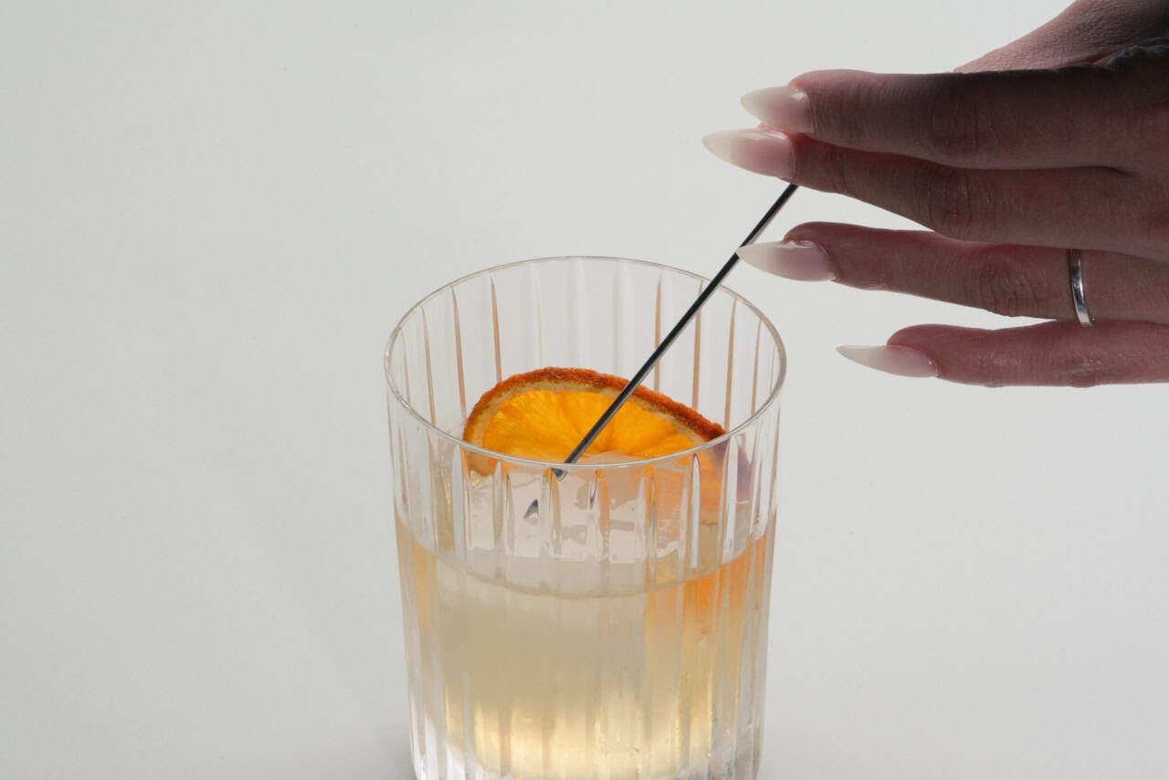 A hand with long nails uses a cocktail pick to arrange a dried orange slice in a clear glass with a light-colored mocktail—perfect for anyone wondering, what is a mocktail and how to serve it stylishly.